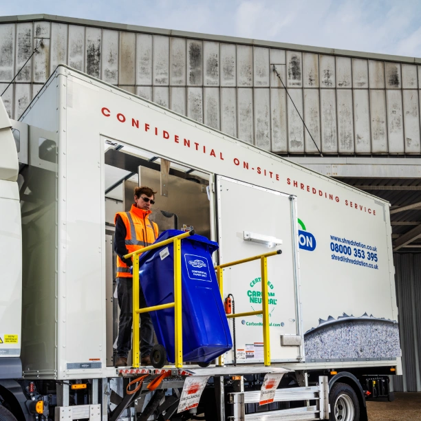 An on-site shredding truck outside a business. A Shred Station operative is loading a blue confidential waste bin into the truck via a side lift.