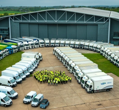 A large fleet of trucks in front of a big grey building. Between the rows of trucks is a large group of people in yellow hi-vis jackets.
