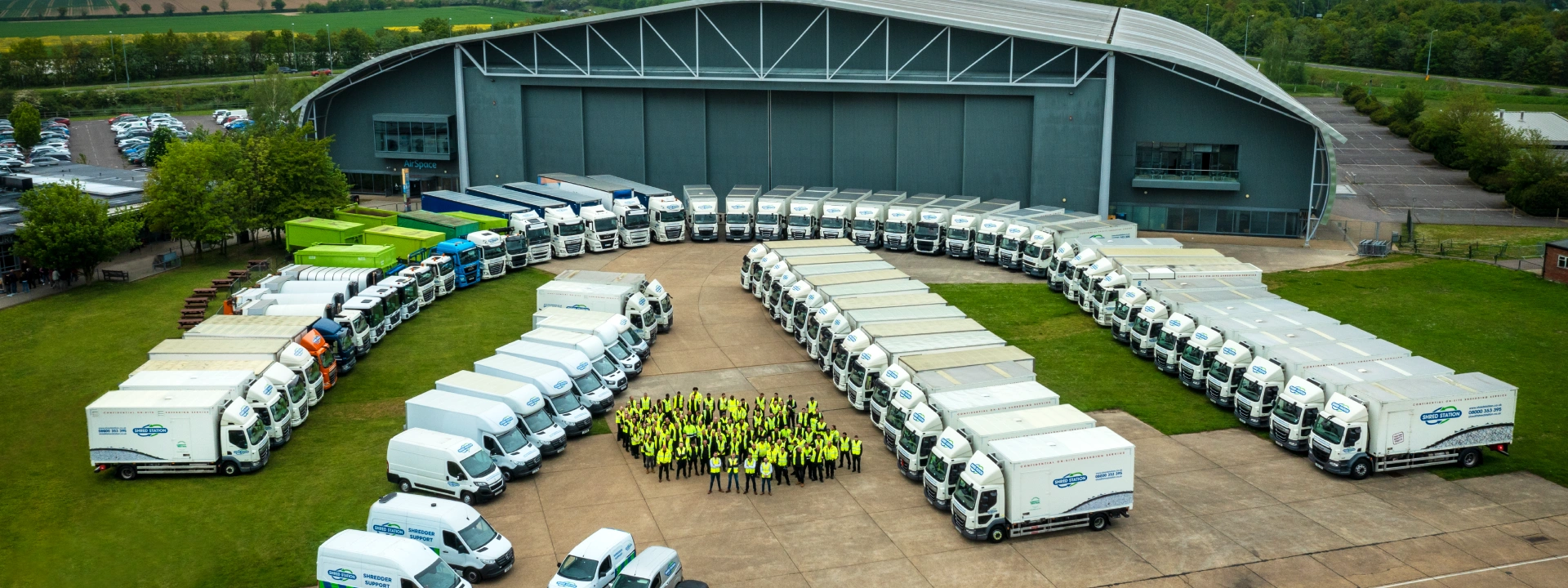 A large fleet of trucks in front of a big grey building. Between the rows of trucks is a large group of people in yellow hi-vis jackets.