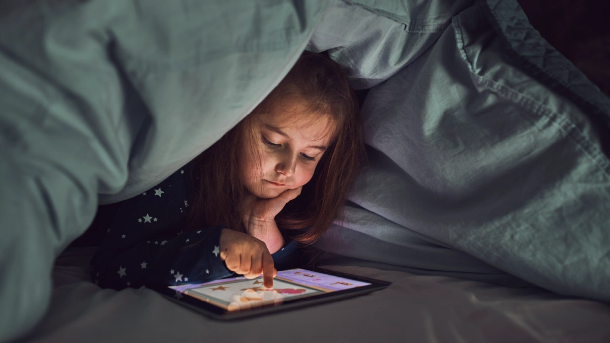 Little girl playing on a tablet under a duvet.