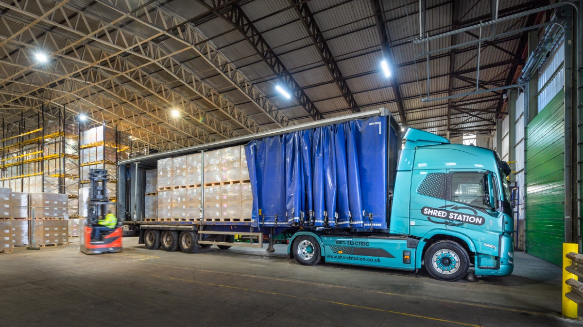 A curtain-side electric HGV articulated lorry unloading pallets of boxes inside a large warehouse.
