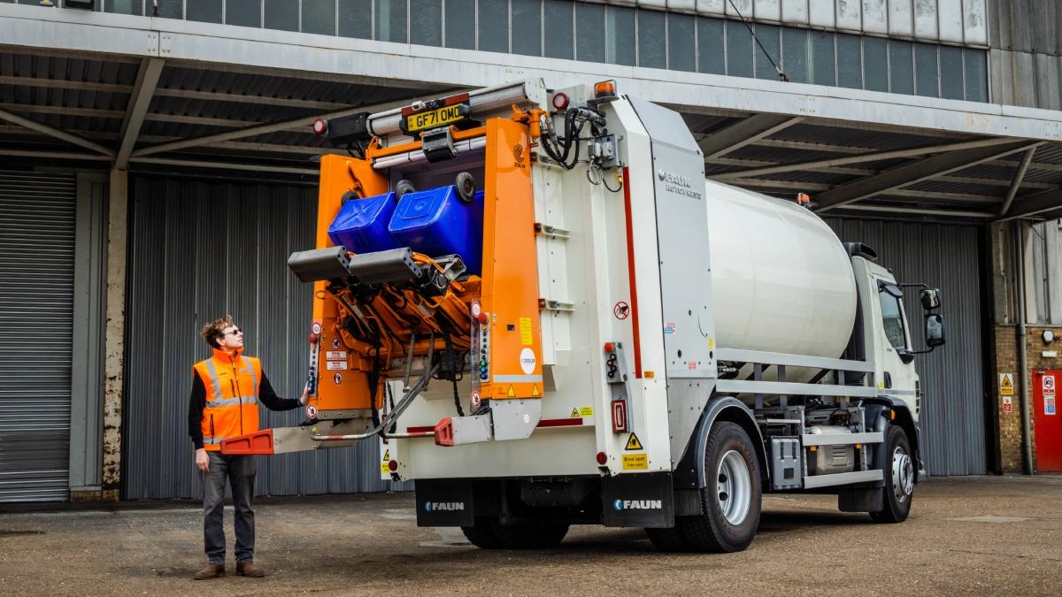 A secure automated vehicle (SAV), parked outside of a warehouse. Two blue confidential waste bins are being electronically tipped into the vehicle by an operative in an orange hi-vis jacket.