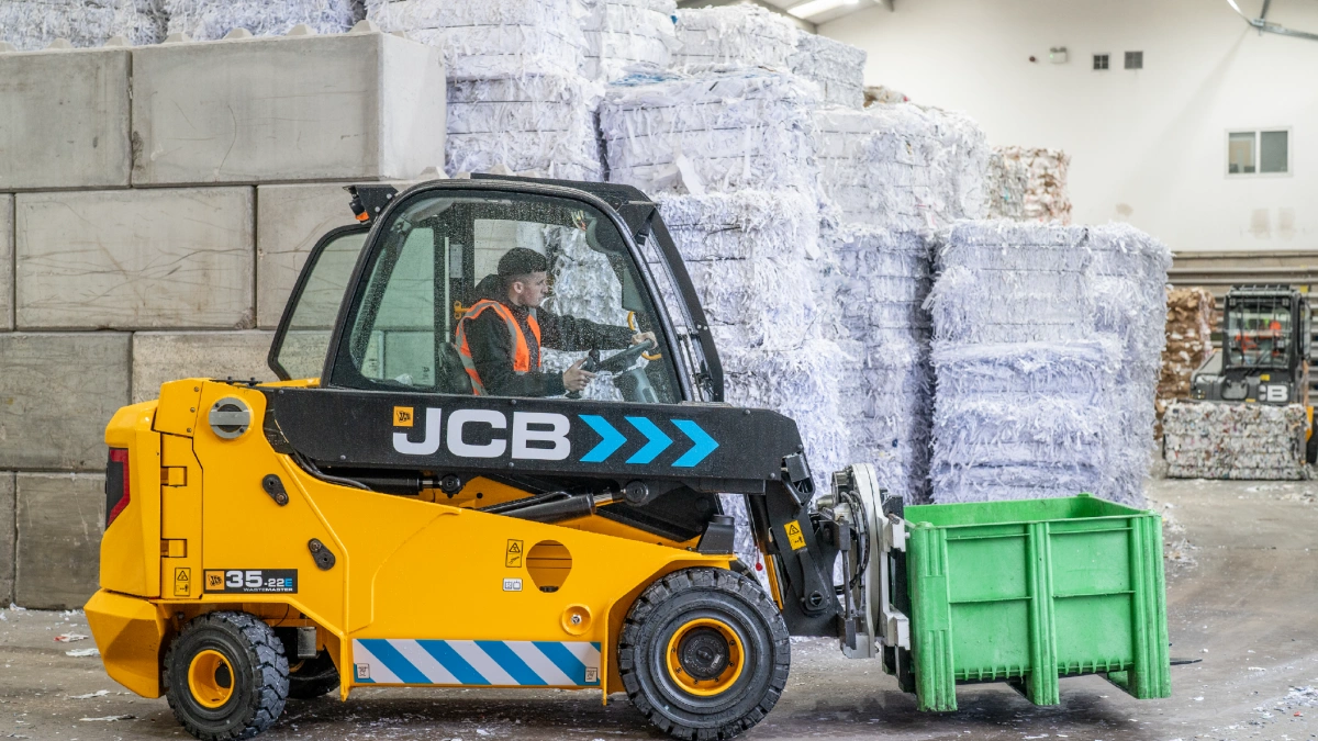 An electric forklift truck with a green dolav on its fork, driving past a large stack of baled plain white paper.