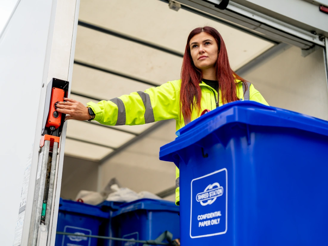 A woman loading a blue confidential waste bin into the back of a truck using a button to raise the tail lift.