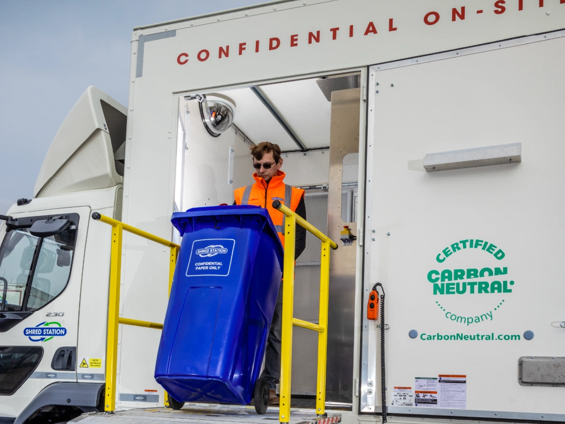 A Shred Station operative wearing dark sunglasses, loading a blue confidential waste bin into an on-site shredding vehicle.