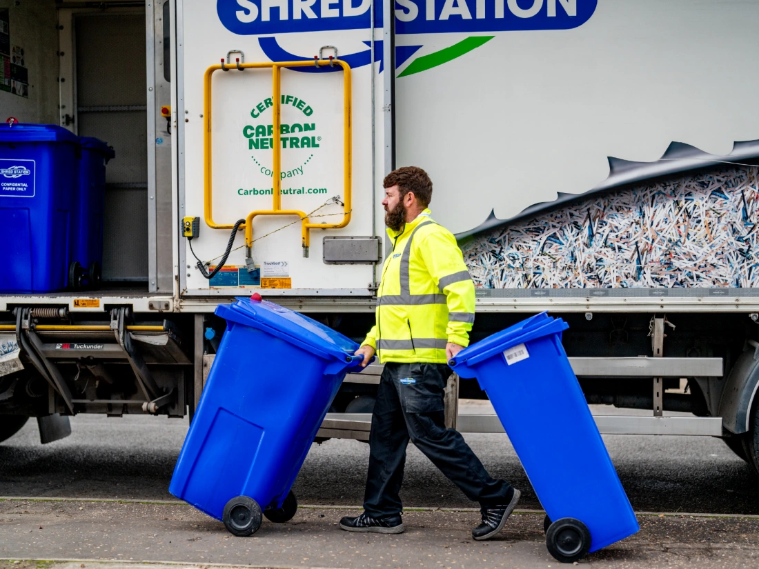 Shred Station operative in full uniform and hi-vis jacket wheeling two blue confidential waste bins towards Shred Station branded truck.