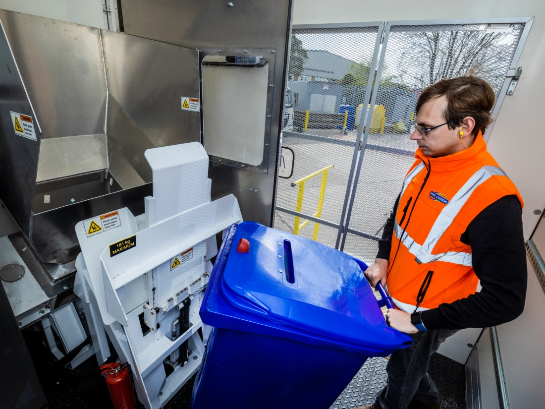 An industrial shredding machine housed within a mobile shredding vehicle. An operative in an orange hi-vis vest is loading a blue confidential waste bin into the machine.