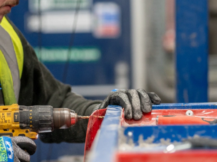 Person in a hi-vis vest unscrewing screws from a blue and red piece of apparatus with a power drill.