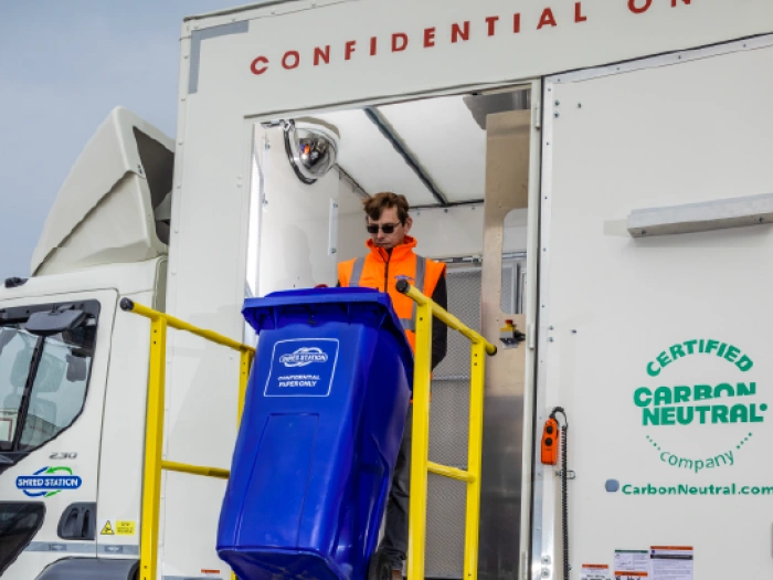 A Shred Station operative wearing dark sunglasses, loading a blue confidential waste bin into an on-site shredding vehicle.