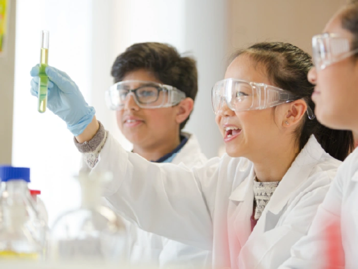 Schoolchildren in lab coats looking at a test tube filled with green liquid.