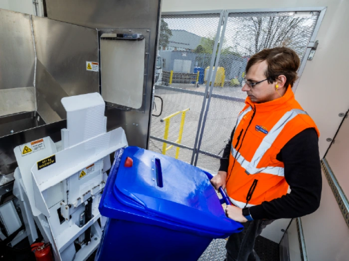 An industrial shredding machine housed within a mobile shredding vehicle. An operative in an orange hi-vis vest is loading a blue confidential waste bin into the machine.
